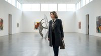 Mature woman with shoulder-length brown hair, Caucasian ethnicity, wearing a black blazer and carrying a brown leather bag, standing in a contemporary art gallery examining a modern metal sculpture. Polished concrete floors and white walls with artwork displayed throughout.