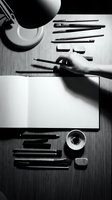 Artist workspace with hand drawing on paper surrounded by professional art supplies and desk lamp in monochrome style for creative design projects.
