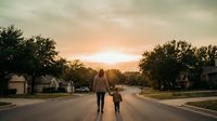Mother and young child walking hand in hand down peaceful suburban street at golden hour sunset with warm sky and residential homes lining quiet neighborhood road.