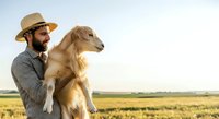 Man with beard wearing straw hat holding golden retriever in rural field landscape. Pet owner with dog outdoors.
