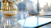 Golden glass candle holder on reflective surface with laptop and pen in soft focus background, creating elegant workspace atmosphere with natural lighting.