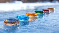 Colorful glass pebbles arranged in line on wet surface with water drops, creating artistic composition with reflections and bokeh effect in natural light.
