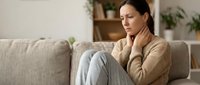 Young woman experiencing neck pain sitting on couch at home, touching sore throat area with hand, health discomfort concept for medical wellness content.