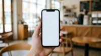 Hand holding smartphone with blank white screen in cozy coffee shop interior, mockup template for mobile app presentation and digital marketing materials.