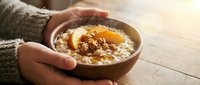Hands holding warm bowl of oatmeal porridge topped with caramelized apples, nuts and cinnamon for breakfast, healthy eating and comfort food concepts.