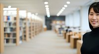 Young Asian woman in professional black attire smiling at camera in modern library with bookshelves and study tables, suitable for education and business concepts.