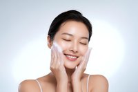 Young Asian woman applying facial cleanser foam with closed eyes and genuine smile, demonstrating skincare routine against white background.