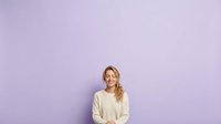 Smiling woman with blonde wavy hair wearing cream sweater against purple background, suitable for portrait, lifestyle, and advertising campaigns.