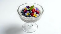 Chia seed pudding in a glass bowl topped with fresh raspberries, blueberries, edible flowers, and mint, isolated on white background.