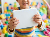 Young child with medium skin tone holding blank tablet device, wearing colorful striped shirt in playful educational environment with toys and learning materials in background.