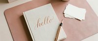 Elegant linen journal with rose gold hello calligraphy, pen, and blank cards on a blush pink desk mat, styled for stationery and lifestyle content.