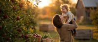 Mother holding toddler child in apple orchard during golden hour sunset creating warm family bonding moment in countryside farm setting.