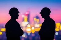 Silhouettes of two construction workers in hard hats against blurred urban skyline with bokeh lights during colorful sunset. Concept of engineering and urban development.