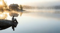 Peaceful morning fishing scene with angler sitting on rocky shore of misty lake at sunrise. Cozy cabin reflects in calm water creating serene outdoor recreation moment.