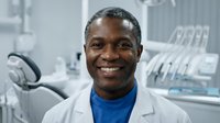 Male dentist of African descent wearing white coat and blue shirt, smiling in modern dental clinic with equipment in background. Portrait for healthcare, medical, and dental practice marketing.