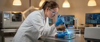 Female scientist in white lab coat and safety glasses conducting research with laboratory equipment in modern scientific facility.