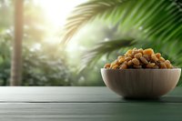 Golden raisins in wooden bowl on rustic table against blurred tropical palm leaves background with sunlight, healthy organic dried fruit snack.