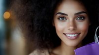 Young African American woman with curly hair smiling warmly while holding purple shopping bags, representing retail therapy and consumer happiness.