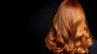 Young woman with luxurious wavy copper red hair styled in soft curls shown from behind against dark background, demonstrating professional hairstyling and coloring technique.