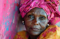 Mature African woman wearing vibrant pink headwrap and yellow traditional dress smiles warmly against pink wall background, radiating joy and cultural pride.