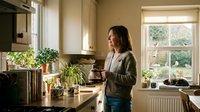 Middle-aged Caucasian woman standing thoughtfully in modern kitchen with houseplants, natural lighting streaming through windows creating warm domestic atmosphere.
