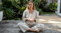 Young woman practicing meditation in lotus position on wooden deck surrounded by tropical plants creating peaceful zen atmosphere for mindfulness and relaxation.