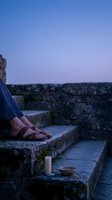 Woman in sandals sitting on ancient stone steps during twilight hour with candle and bowl creating peaceful meditation atmosphere for wellness content.