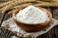 White flour in wooden bowl on rustic linen napkin, wheat ears on dark wooden table background, natural baking ingredients composition.