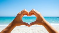 Hands forming heart shape against turquoise ocean and white sand beach on sunny day. Summer vacation love concept for romantic travel destinations.