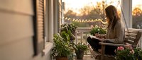 Young woman relaxing on cozy balcony with plants during golden hour sunset, enjoying peaceful evening moment in urban garden setting.