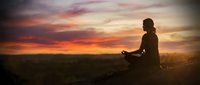 Woman practicing yoga meditation at sunset with dramatic colorful sky and peaceful landscape creating serene wellness atmosphere for mindfulness and relaxation.