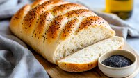 Freshly baked poppy seed bread loaf sliced on wooden cutting board with black sesame seeds in ceramic bowl, soft white crumb texture and golden crust.