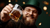 Cheerful bearded man in glasses and bowler hat raising beer mug with amber ale against blurred bokeh lights background, celebrating at pub or party.