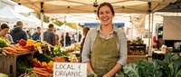 Young Caucasian female farmer smiling at organic farmers market stand with fresh vegetables and local produce display for healthy eating and sustainable agriculture.
