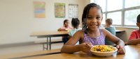 Happy African American elementary school girl enjoying healthy lunch meal in bright classroom cafeteria with classmates in background.