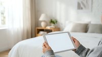 Person holding tablet with blank screen in bright bedroom with white bedding, wooden nightstand, lamp, and window with curtains, suitable for technology, home automation, and lifestyle marketing.