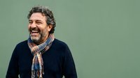 Middle-aged man with curly dark hair and beard, wearing navy sweater and patterned scarf, smiling against green background. Portrait for lifestyle, fashion, advertising.