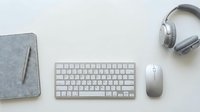 Minimalist workspace flat lay with wireless keyboard, mouse, headphones, and felt notebook on white background.