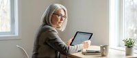 Senior businesswoman with gray hair and glasses working on tablet computer at modern office desk near window for professional consulting services.
