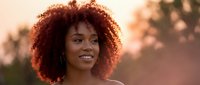 Young African American woman with curly afro hair smiling warmly during golden hour sunset, natural beauty portrait for lifestyle and wellness content.