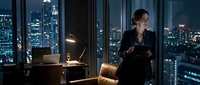 Young professional woman in business attire standing in modern high-rise office at night, overlooking illuminated cityscape through floor-to-ceiling windows, suitable for corporate and business concepts.
