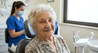 Happy elderly Caucasian woman smiling in dental office chair with female dentist in blue scrubs and mask working in background during routine checkup.