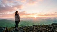 Woman with long brown hair wearing casual athletic clothing standing on rocky mountain peak overlooking green valley landscape at golden hour sunset. Outdoor adventure and nature exploration concept.