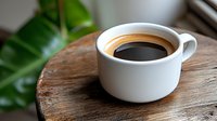 Fresh black coffee in white ceramic cup on rustic wooden surface with green plant leaves in background, morning beverage photography for cafe menu or blog.
