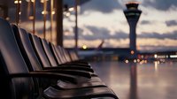 Empty airport terminal seating area at sunset with control tower silhouette visible through large windows, dramatic sky and bokeh lights create atmospheric mood.
