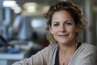 Confident middle-aged Caucasian businesswoman with curly hair smiling warmly at camera in modern office environment, wearing casual beige sweater.