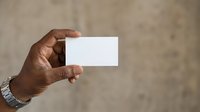 Male hand with dark skin tone holding blank white business card against neutral beige wall background, suitable for branding, corporate identity, and marketing mockup projects.