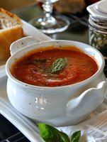 Fresh homemade tomato soup garnished with basil leaves served in white ceramic bowl, accompanied by crusty bread. Rich red color and herbs create appetizing presentation.