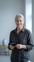 Confident senior Caucasian woman with gray hair holding smartphone in modern office, smiling at camera wearing casual dark shirt for business communication.