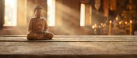 Bronze Buddha statue in meditation pose on wooden surface with warm golden sunlight streaming through windows creating peaceful spiritual atmosphere.